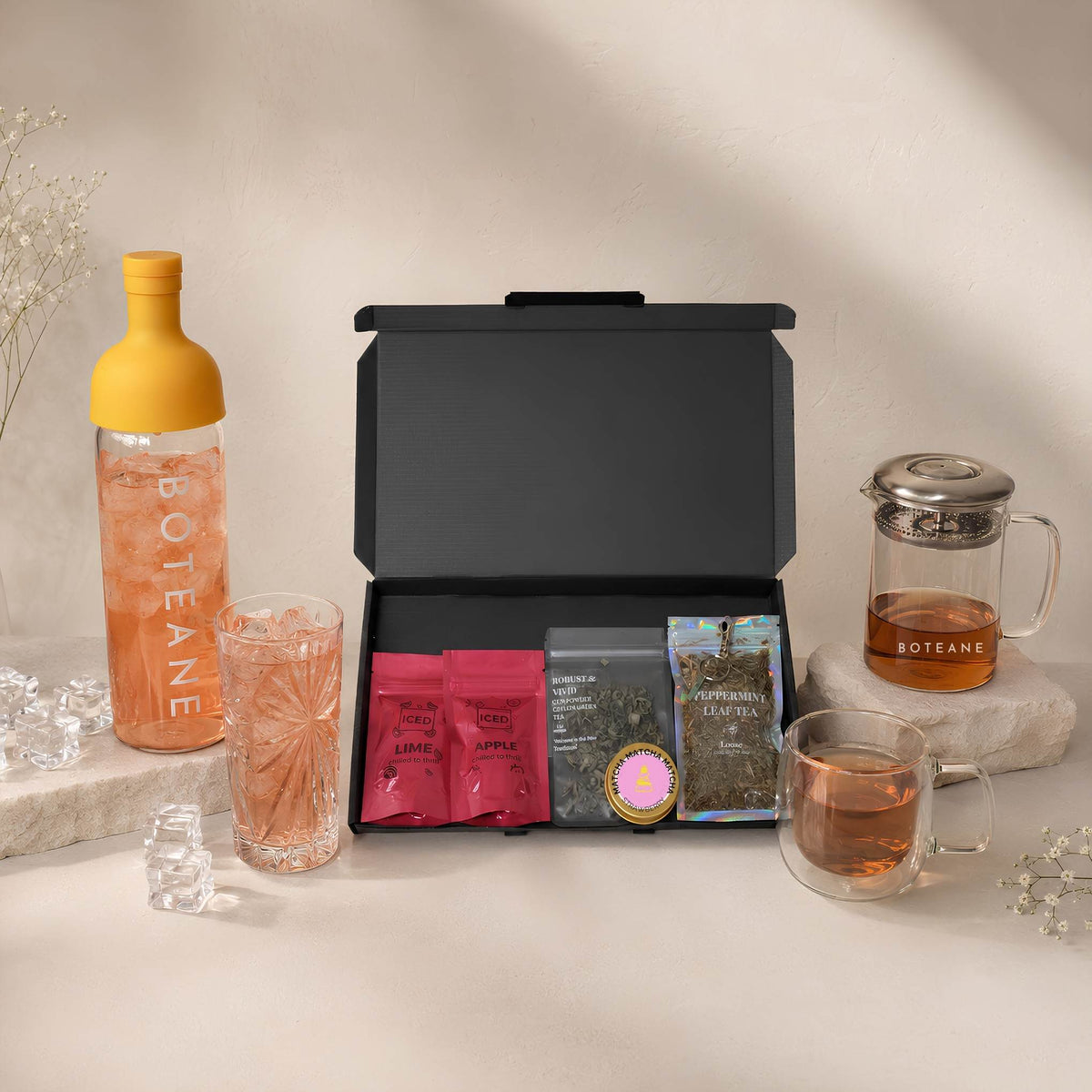 An open black Boteane gift box of loose leaf tea pouches beside a cold brew bottle, a glass teapot and two filled glasses