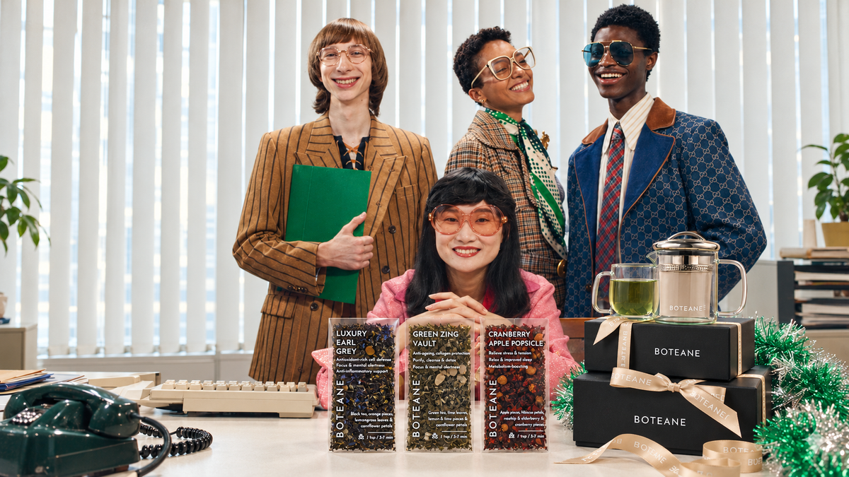 Four colleagues in a retro office with Boteane tea pouches, a glass teapot and stacked black Boteane gift boxes tied with cream ribbon