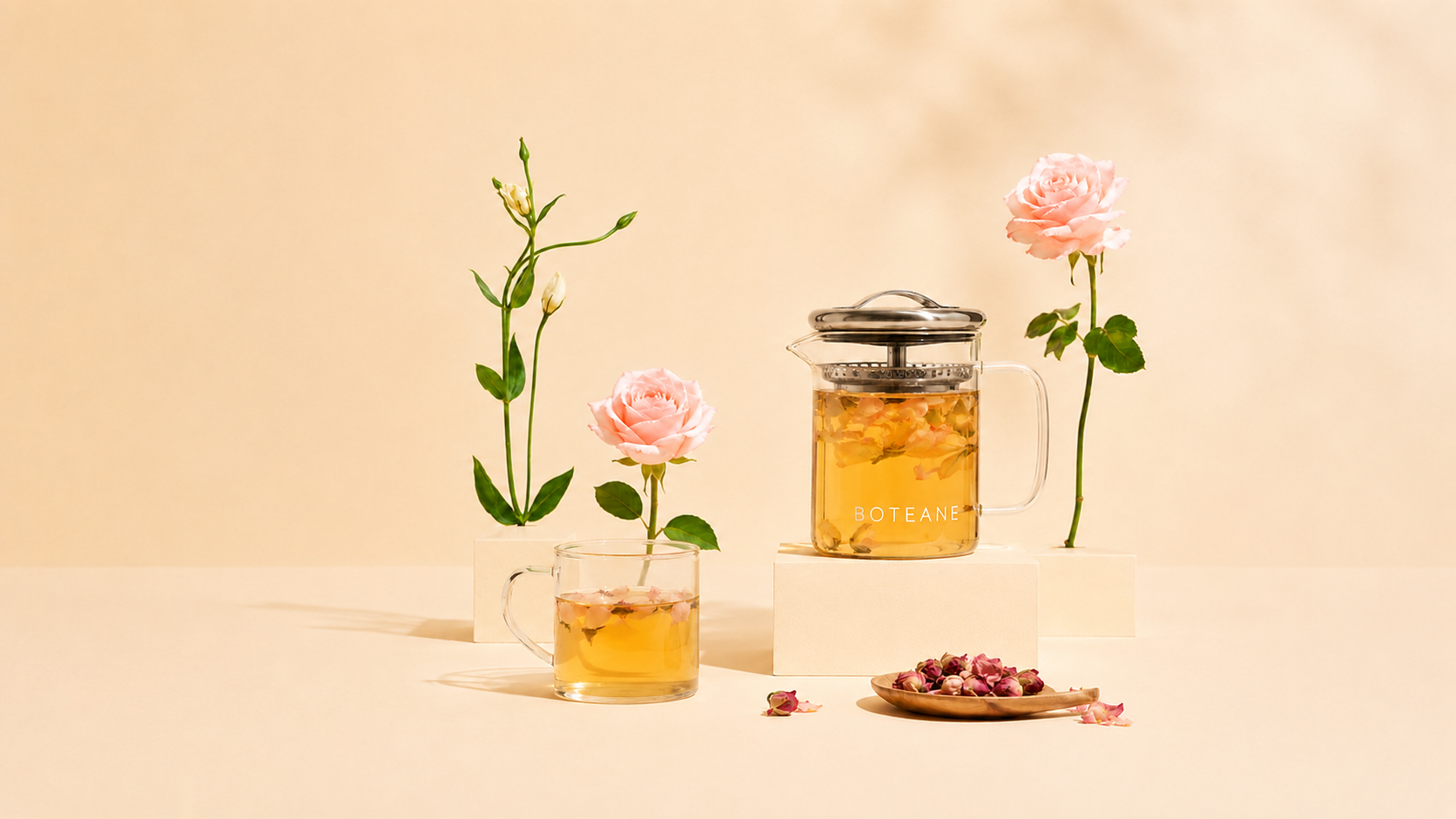 A glass teapot and cup of amber tea on a white pedestal beside pink roses, on a warm beige background