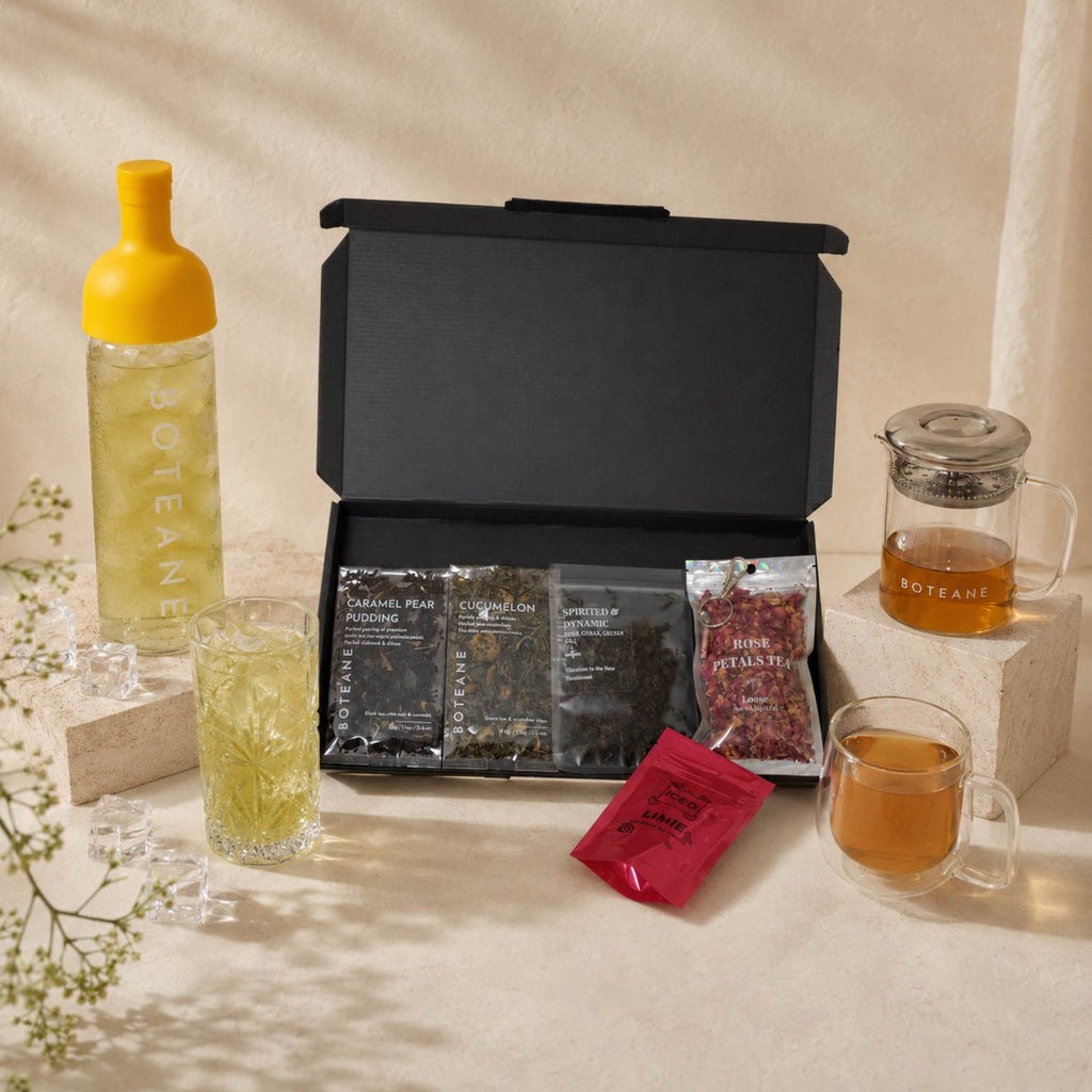 A tea gift set arranged on a beige surface with a black presentation box open to show several tea packets, a yellow-topped bottle, a glass of pale tea, a glass teapot, and a glass cup of amber tea. No people or real environment are present, so this is Product/Packaging rather than Lifestyle.