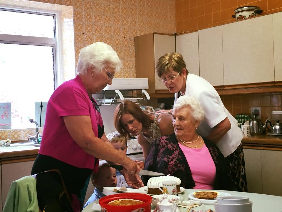 Family gathered in a kitchen around a table, serving cake and tea together