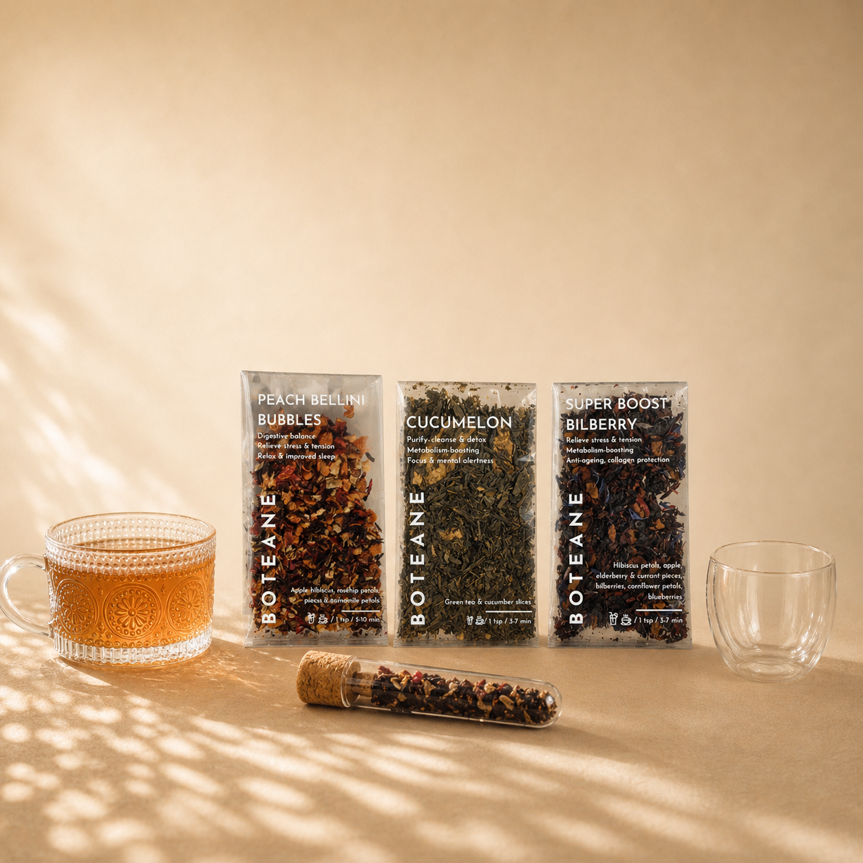 Three Boteane tea packages on a surface with a glass of tea and a clear glass.